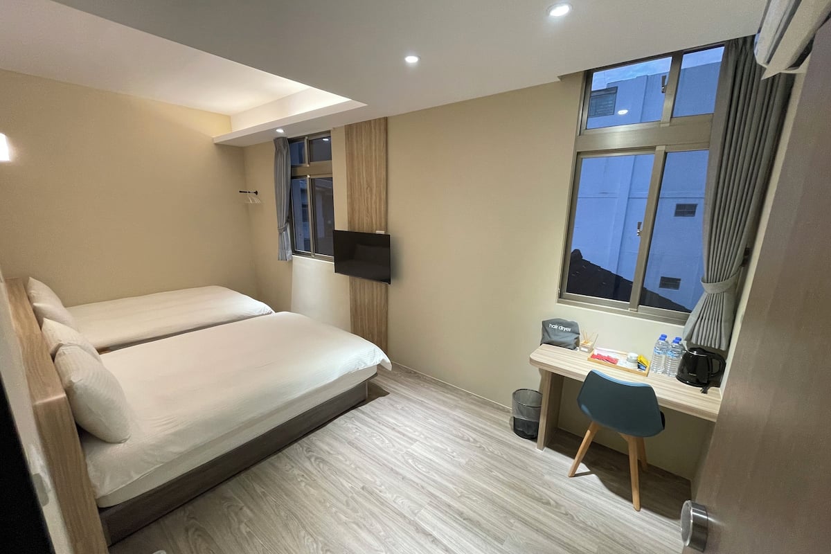 A hotel room with a bed, a desk, a chair, a TV, and a window with a view of buildings.