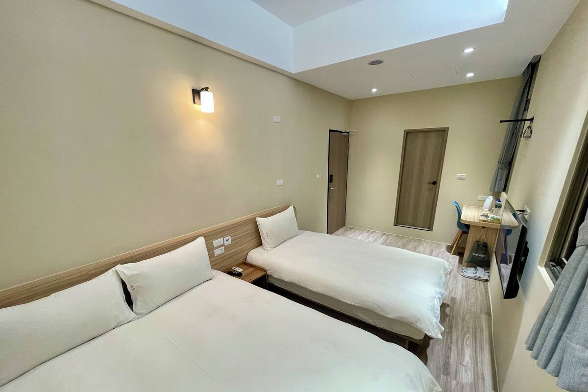 A hotel room with two beds, a desk with a chair, a small table, a mirror, and a window with curtains.