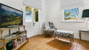 Ein Zimmer mit Holzboden, einem Fernseher auf einem Holzschrank, einem Stuhl, einer Bank und einem Fenster mit Blick ins Grüne.