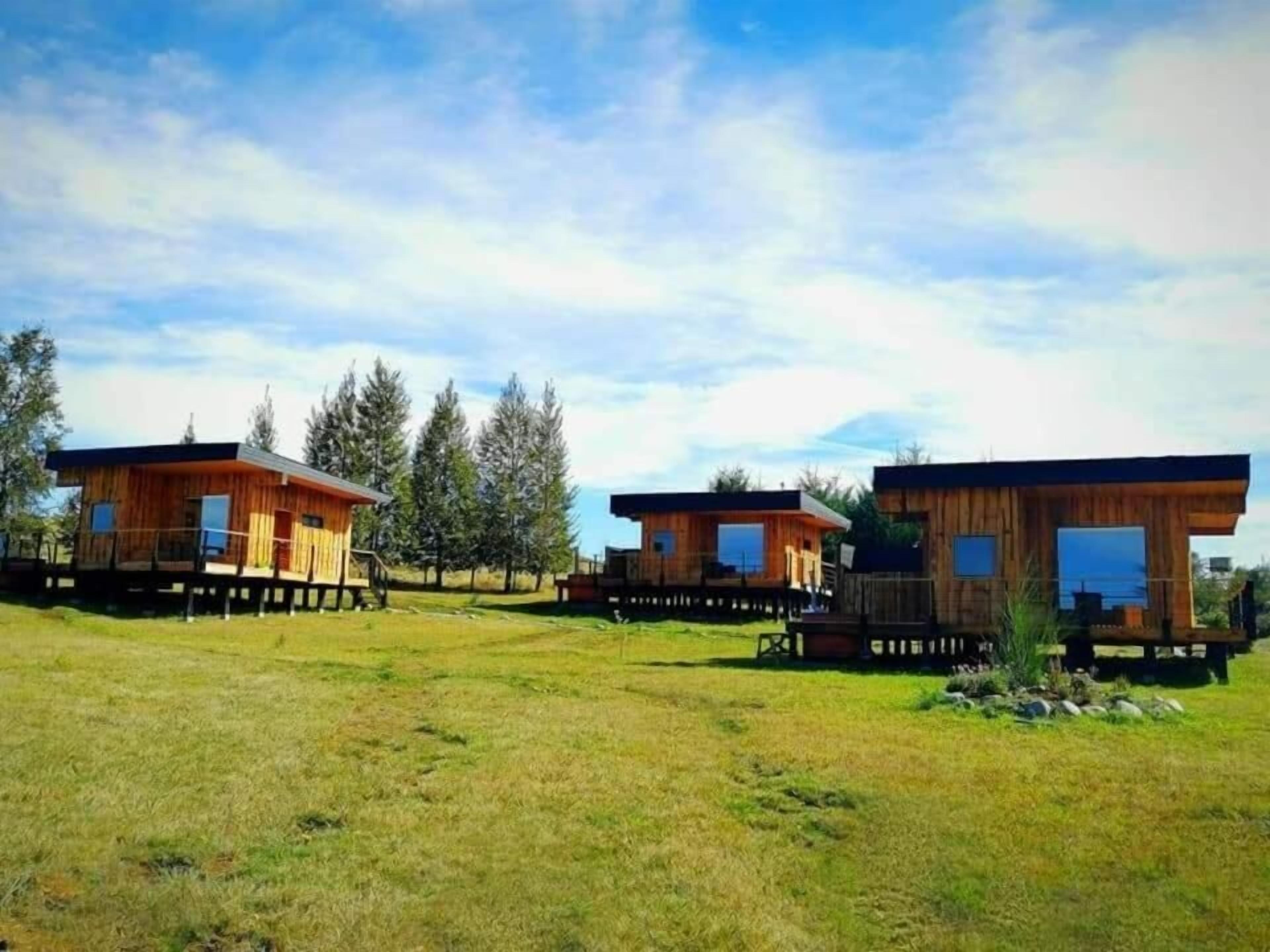 Trois cabanes en bois dotées d’une terrasse, entourées d’un champ verdoyant et d’arbres, sous un ciel partiellement nuageux.