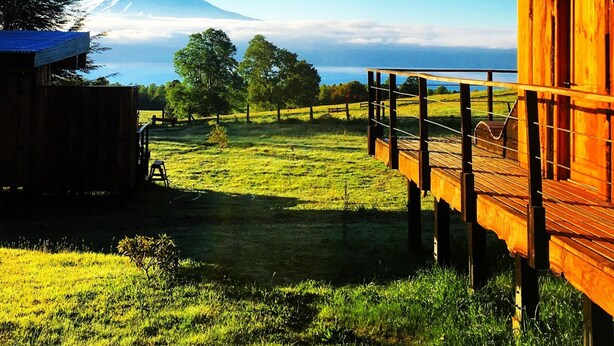 Una cabaña de madera con terraza que da a un campo cubierto de hierba y una montaña al fondo.