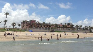 Una playa con un edificio grande al fondo, palmeras y gente relajándose en la arena.