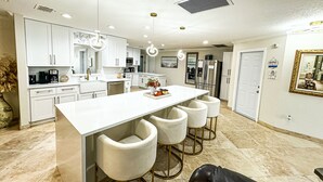 Una cocina moderna con armarios blancos, una isla central con taburetes altos, electrodomésticos de acero inoxidable e iluminación empotrada en el techo.