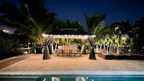 Un área junto a la piscina con zona de estar cubierta, sillones de descanso y una pérgola con luces colgantes.