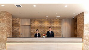 A reception desk with two staff members, a brick-patterned wall, and a marble-tiled floor. - Amagasaki Plaza Hotel Hanshin Amagasaki (Amagasaki)