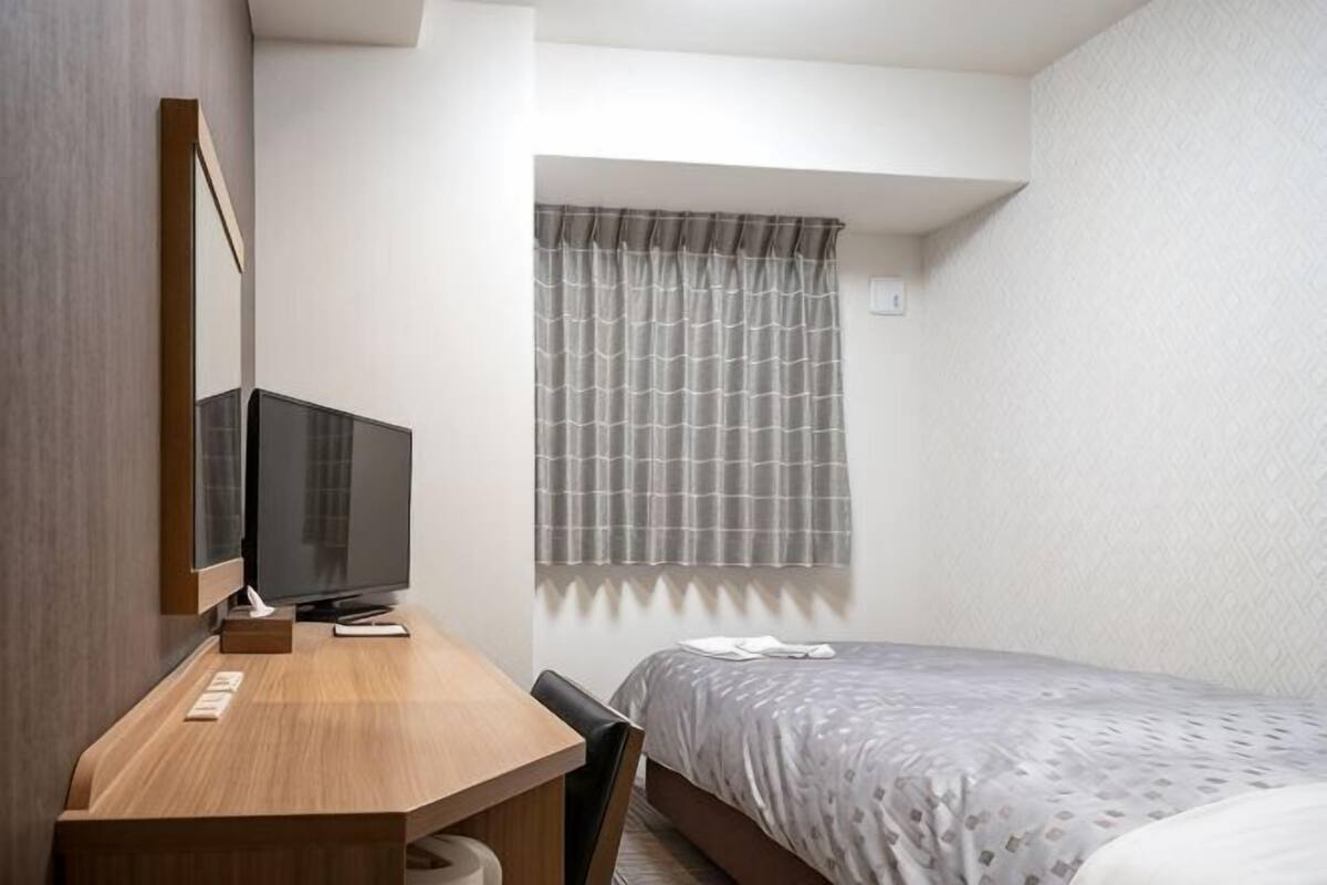 Ein kleines Hotelzimmer mit Bett, Schreibtisch mit Fernseher, Sessel und einem Fenster mit Vorhängen.