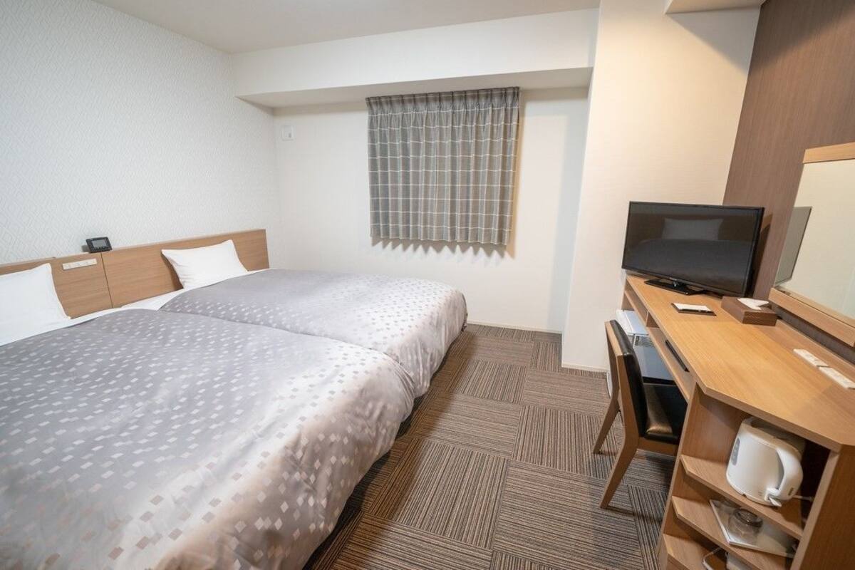 Ein Hotelzimmer mit einem Bett, einem Schreibtisch mit Computer, einem Fernseher und einem Fenster mit Vorhängen.