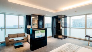 Une chambre d’hôtel moderne dotée d’une grande télévision, d’un canapé, d’une petite table et offrant une vue sur la ville grâce à de grandes fenêtres.