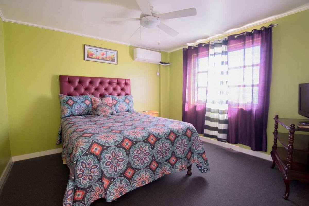 A bedroom with a tufted headboard, a bed with a patterned bedspread, a ceiling fan, a window with purple curtains, and a television on a wooden stand.