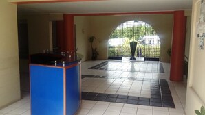 Una hall con una reception blu, pavimento piastrellato e vista attraverso una porta in vetro su una zona verde esterna.