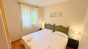 A neatly made bed with white linens and two pillows, a wooden headboard, a nightstand with a lamp, and a window with sheer curtains.
