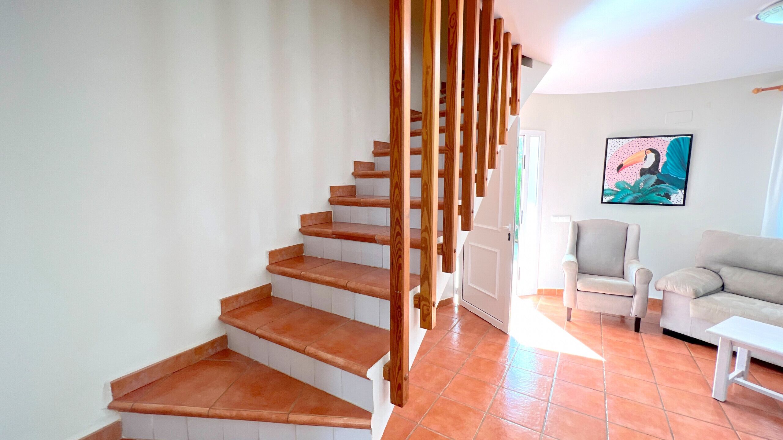 Eine Treppe mit hölzernen Geländern und Terrakottafliesen führt zu einer offenen Tür. Im Wohnbereich befinden sich ein beiges Sofa und ein weißer Tisch.