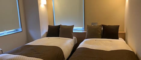 A hotel room with two beds, each with a patterned bedspread and pillows, a window with blinds, and a wall-mounted light fixture.