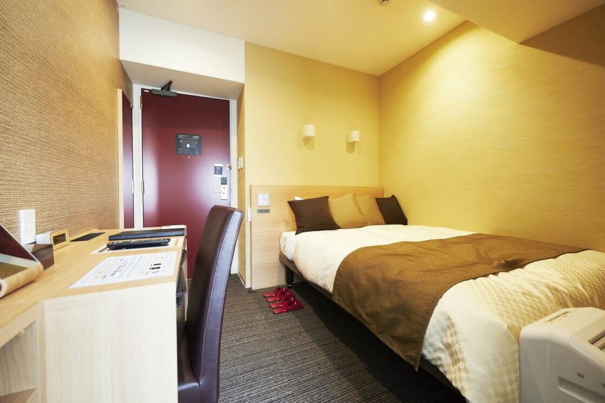 Habitación de hotel con cama, escritorio, silla y una pequeña mesa con computadora y documentos.