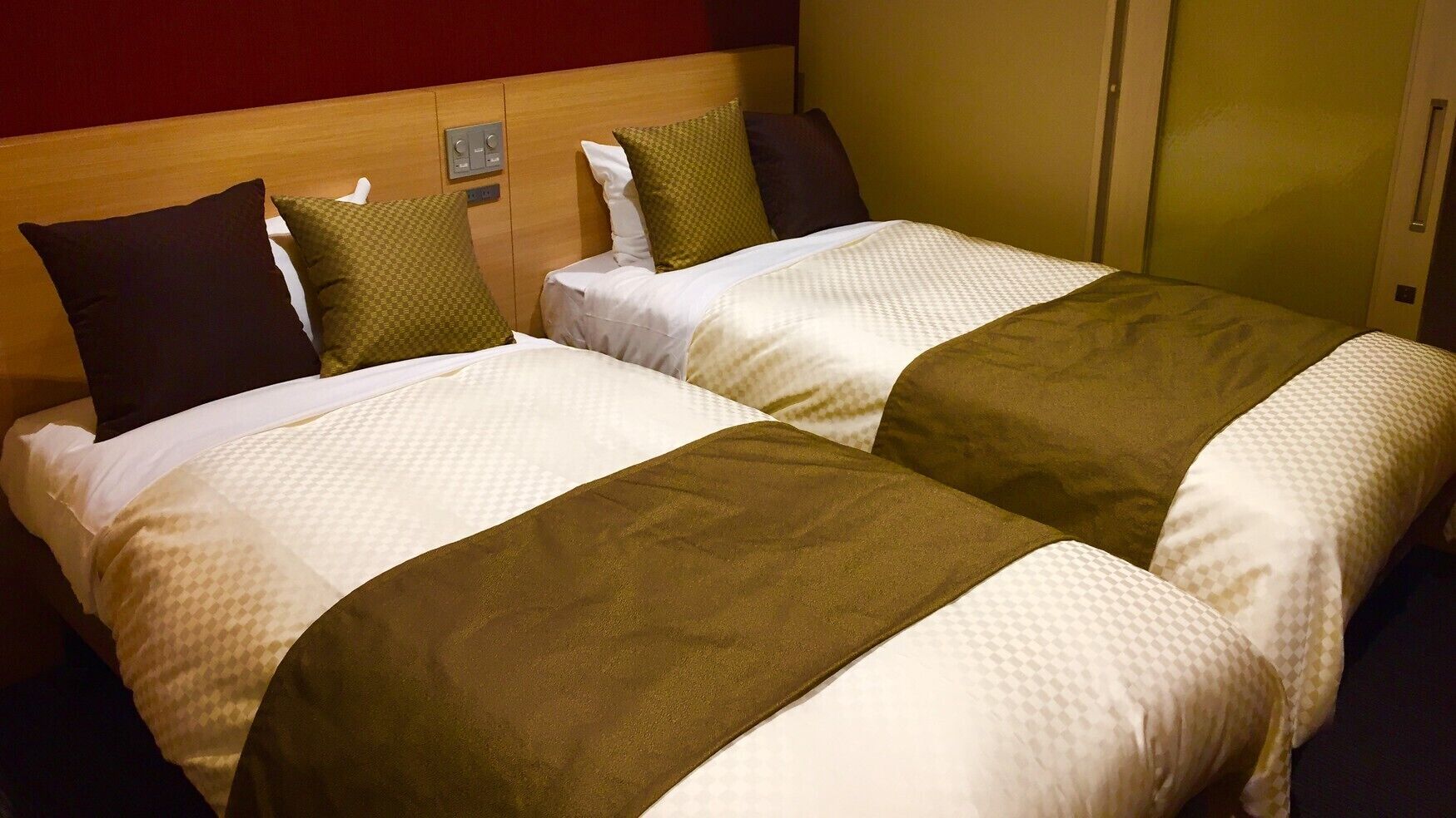 Duas camas com roupa de cama branca e mantas marrons, uma cabeceira de madeira e um painel de controle na parede.