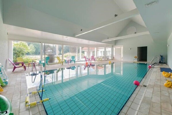 An indoor swimming pool with a tiled floor, surrounded by colorful chairs and a glass enclosure.