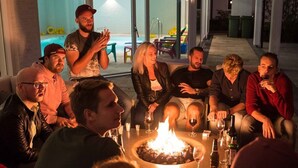 Un groupe de personnes est assis autour d’un foyer, des boissons à la main, dans un espace intérieur bien éclairé. On aperçoit une piscine à l’arrière-plan.