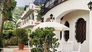 Une cour avec un arbre en pot, des chaises blanches et un parasol blanc.