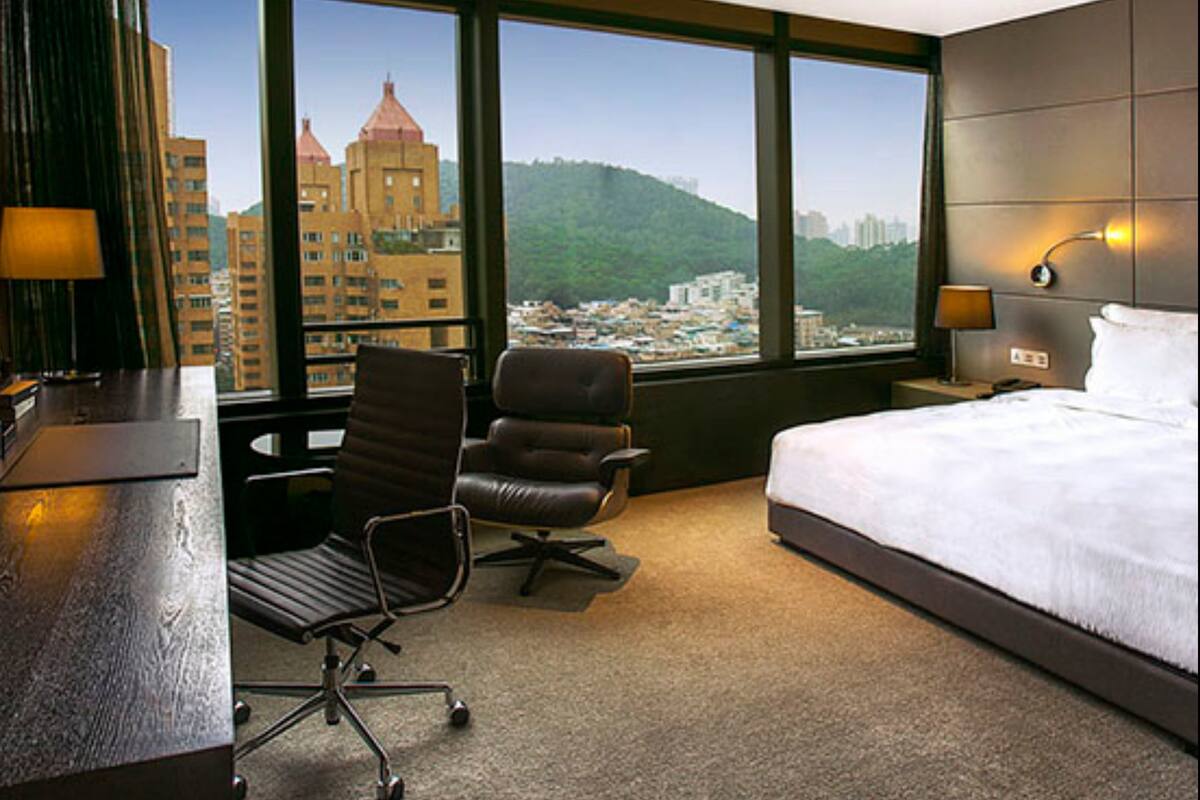 A hotel room with a large bed, a desk, a chair, and a view of a cityscape through large windows.
