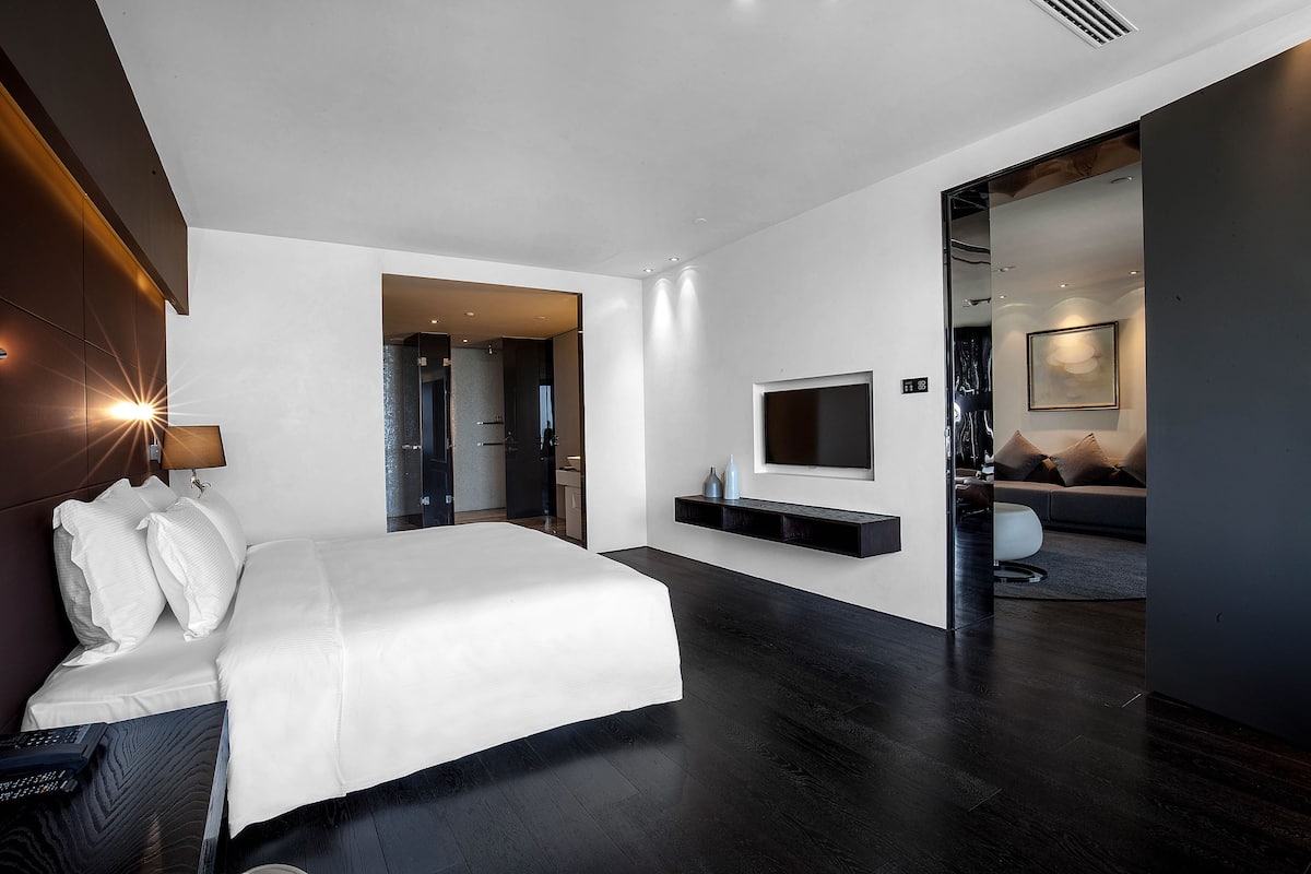 A modern hotel room with a large bed, a flat-screen TV mounted on the wall, and a dark wood floor.
