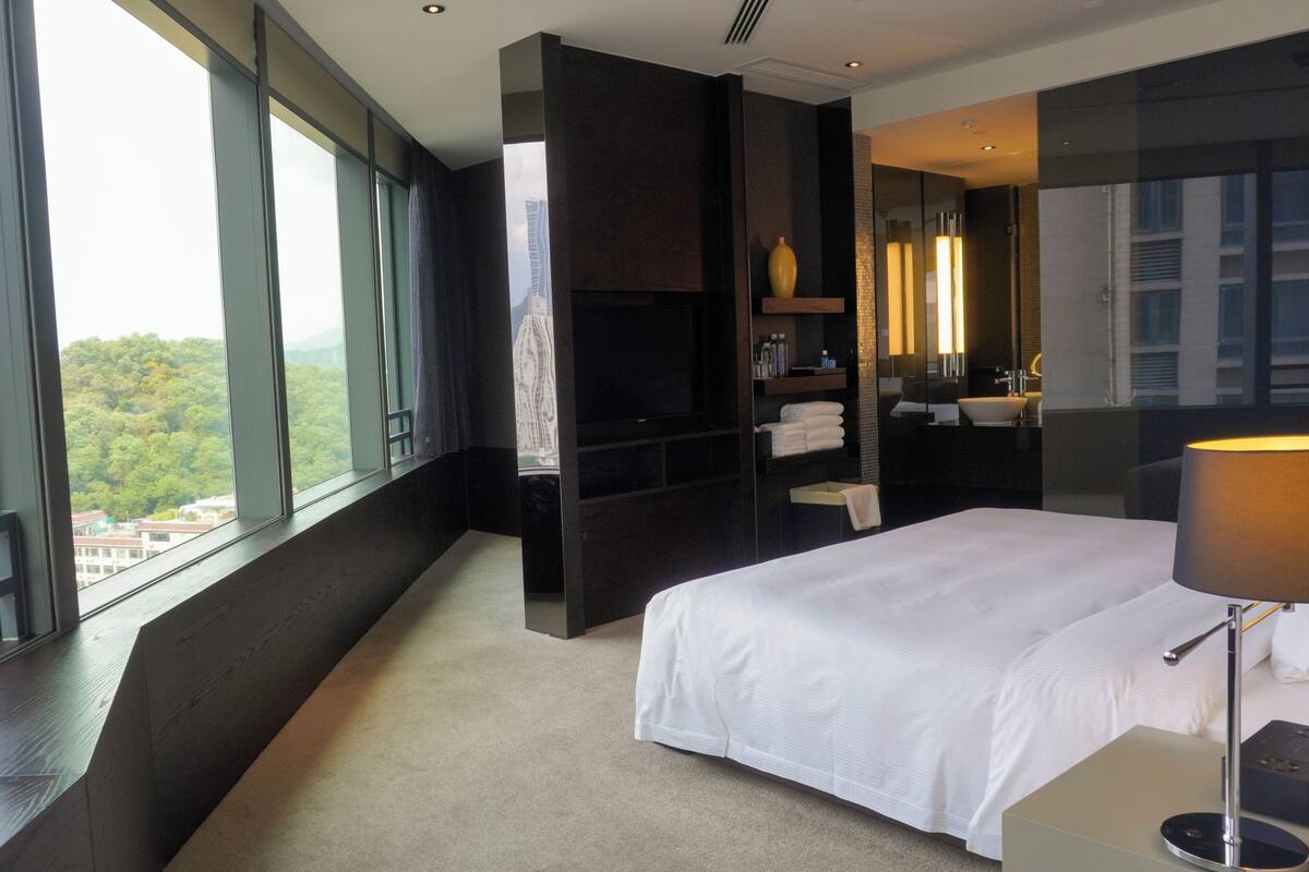 A modern hotel room with a large bed, a TV, a bathroom with a mirror, and a view of a cityscape.