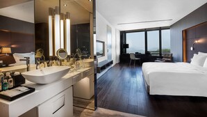 Ein modernes Hotelzimmer mit einem großen Bett, einer separaten Sitzecke, einem großen Fenster mit Blick und einem Badezimmer mit großem Spiegel und Waschbecken.