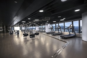 Un gimnasio moderno con cintas de correr, máquinas de pesas y bancos, con amplias ventanas y vistas a la ciudad.