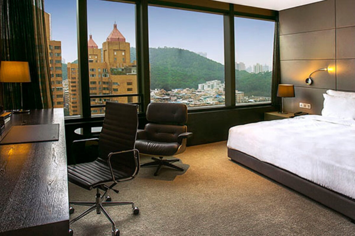 Habitación de hotel con una cama grande, un escritorio, una silla y vistas a la ciudad a través de amplias ventanas.
