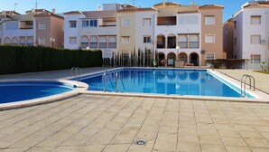 A swimming pool area with tiled flooring, a clear blue pool, and a building with multiple balconies and windows in the background. - Perfectly located, refurbished apartment in a gated complex with pool and garden (Torrevieja)