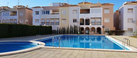 A swimming pool area with tiled flooring, a clear blue pool, and a building with multiple balconies and windows in the background.