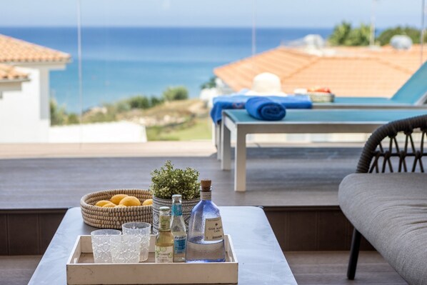 A tray with gin, tonic, and other beverages, a basket of lemons, and a potted plant on a table with a view of the sea and a lounge area.