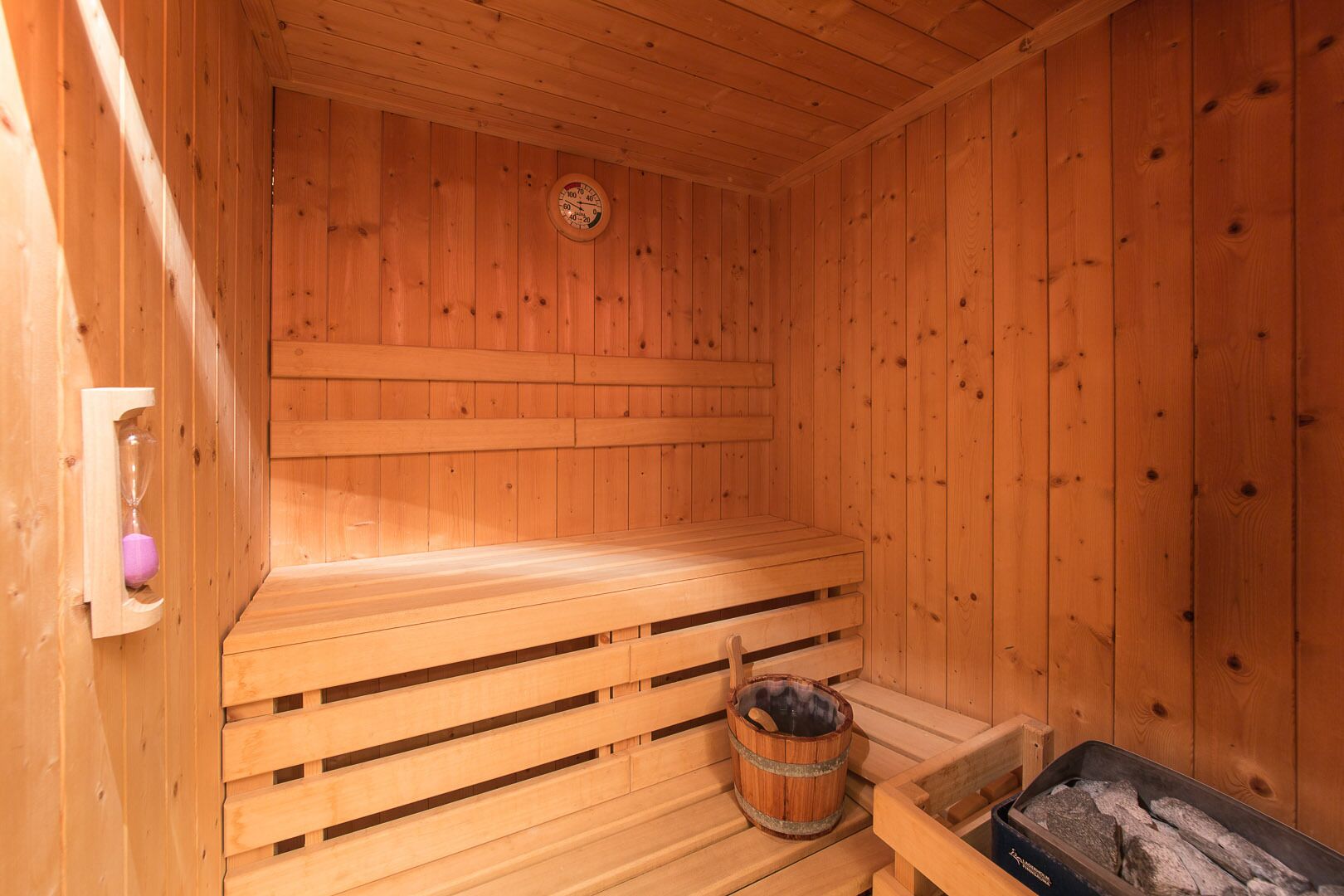 A wooden sauna with benches, a wooden bucket, and a thermometer on the wall.