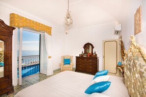 A bedroom with a bed, a dresser, a mirror, a chandelier, and a view of the sea.
