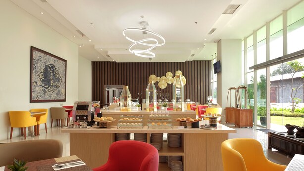 A modern breakfast bar with a variety of pastries and beverages, surrounded by vibrant yellow and red chairs, and large windows offering a view of the outdoors. - Hotel Horison Nindya Semarang (Semarang)