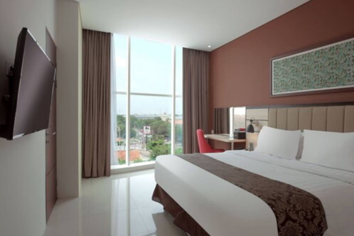 A hotel room with a large bed, a desk, a chair, a TV, and a window with a city view.