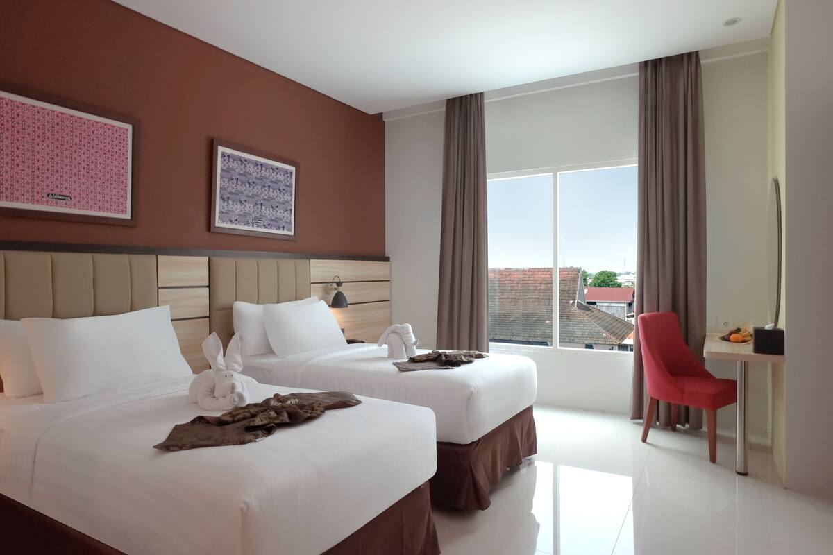 A hotel room with two beds, a desk, a chair, and large windows with curtains.