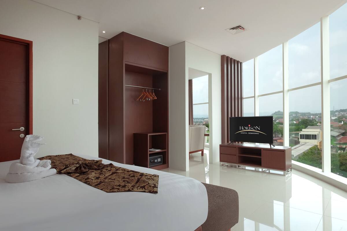 A modern hotel room with a large bed, a wardrobe, a television, and a view of the city.