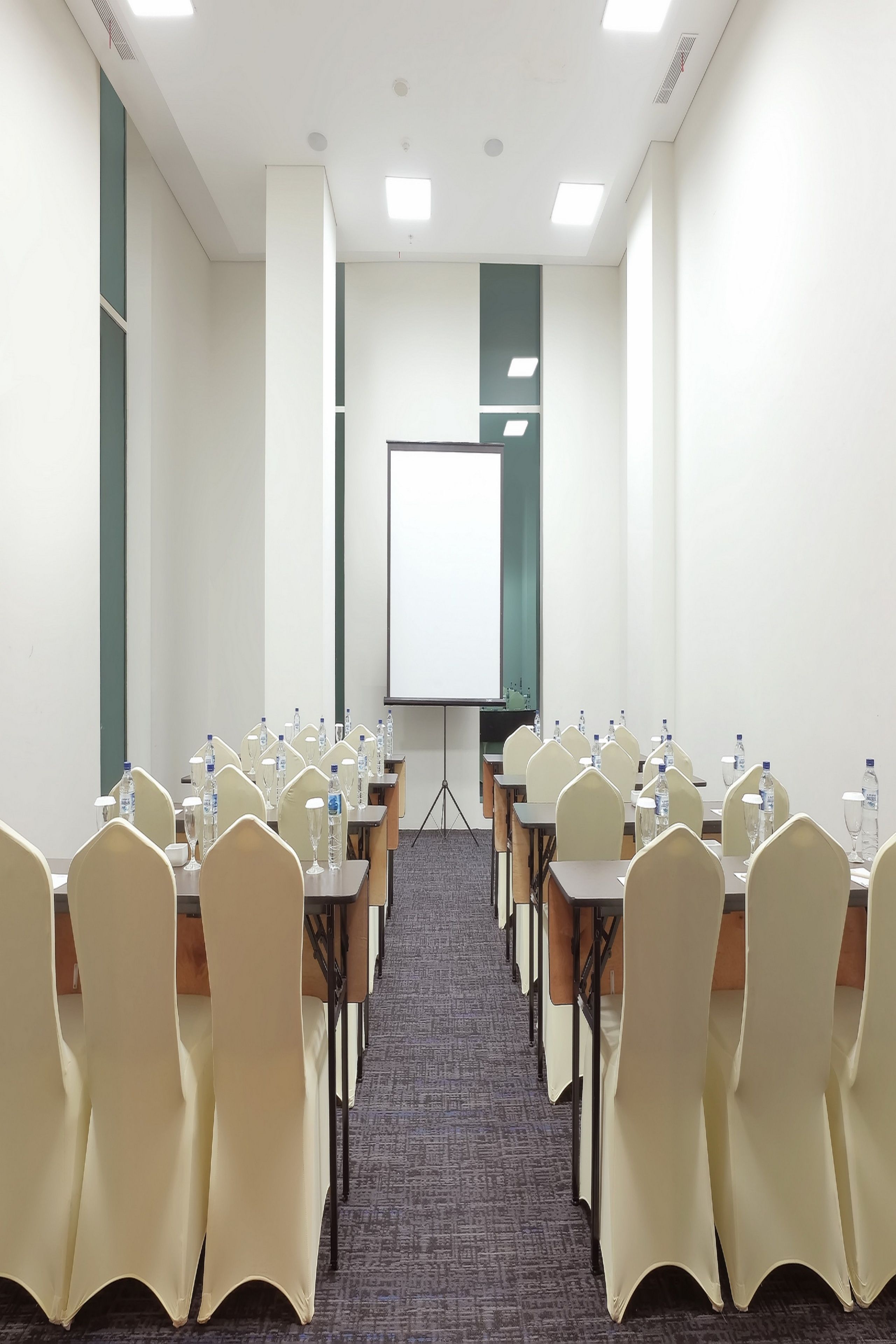 A conference room with rows of tables and chairs, a projector screen, and water bottles on each table.