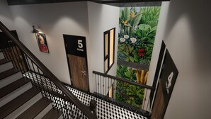 Una escalera con piso a cuadros blanco y negro, barandal de madera y pared con números de habitación y un mural tropical.