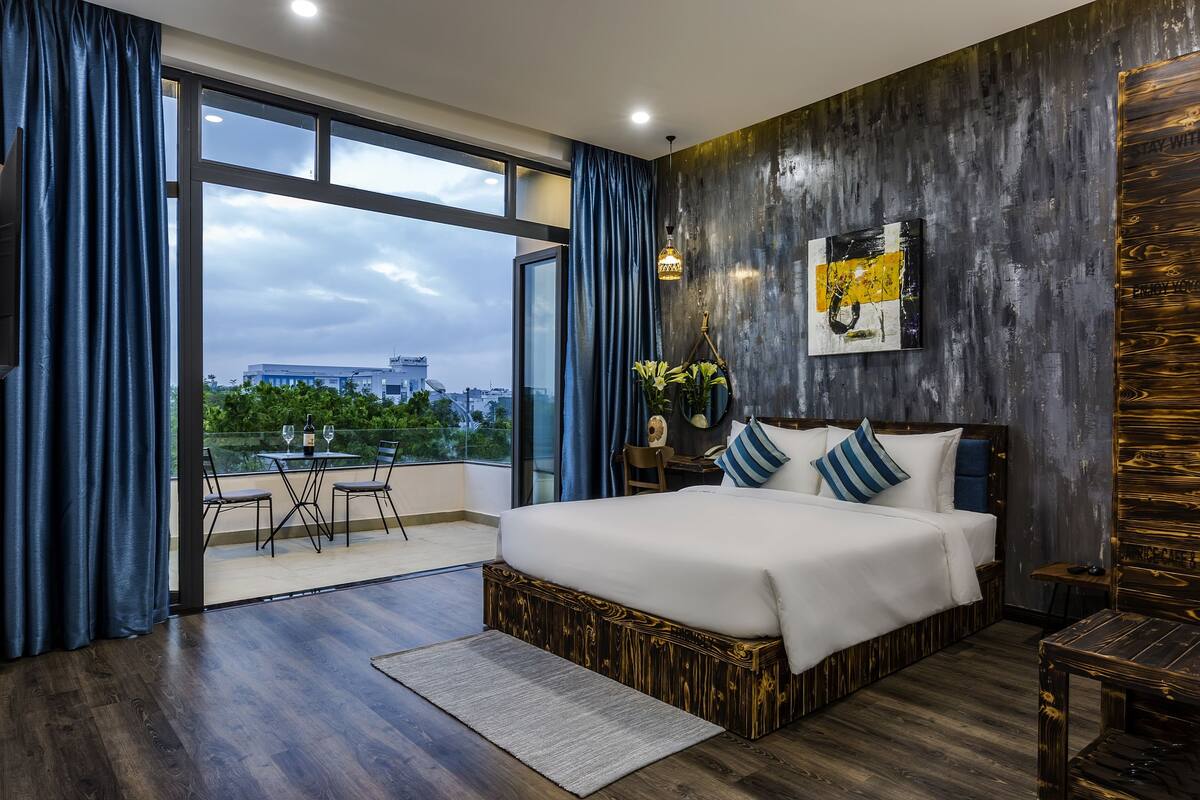 A modern bedroom with a large bed, a balcony with a dining table, and a city view.