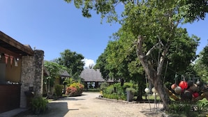 A gravel driveway leading to a rustic building with a stone facade and a thatched roof structure in the background. - Vanuatu Uncharted Sport Fishing Lounge (Mele)