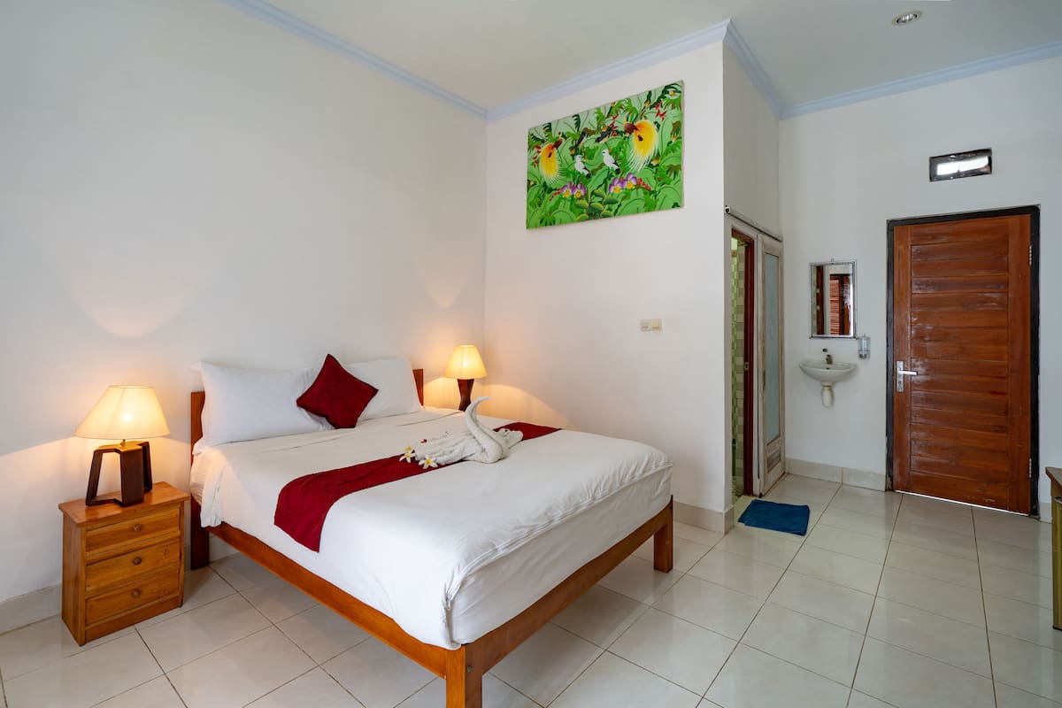 A neatly made bed with a red throw blanket, a wooden nightstand with a lamp, a wall-mounted painting, a bathroom with a sink and mirror, and a wooden door.