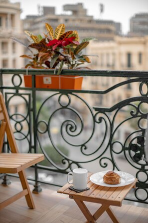 A balcony with a wooden chair, a small table with a plate of food and a cup, and a potted plant.