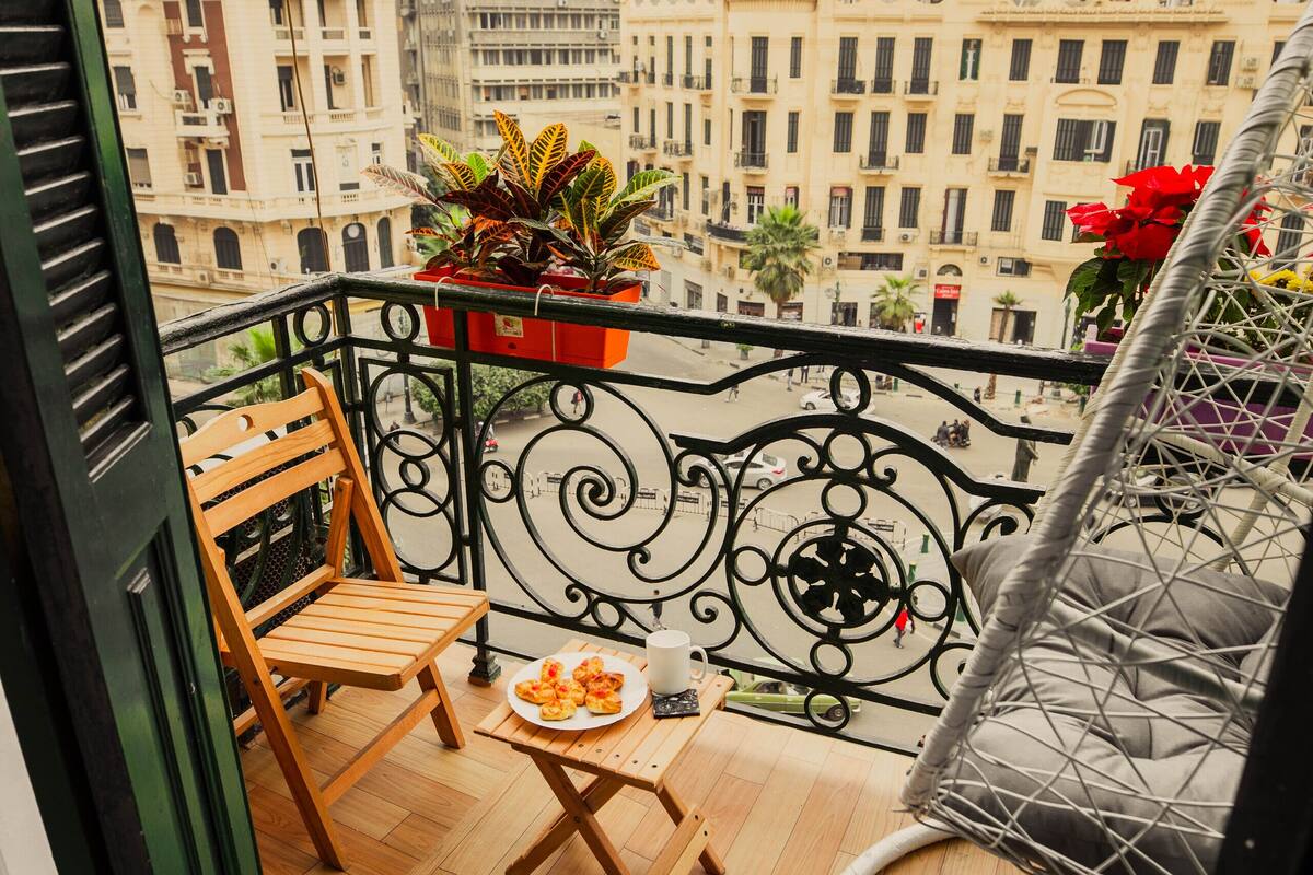 Un balcón con una silla de madera, una mesa pequeña con un plato de comida, una planta en maceta y vistas de edificios de la ciudad.