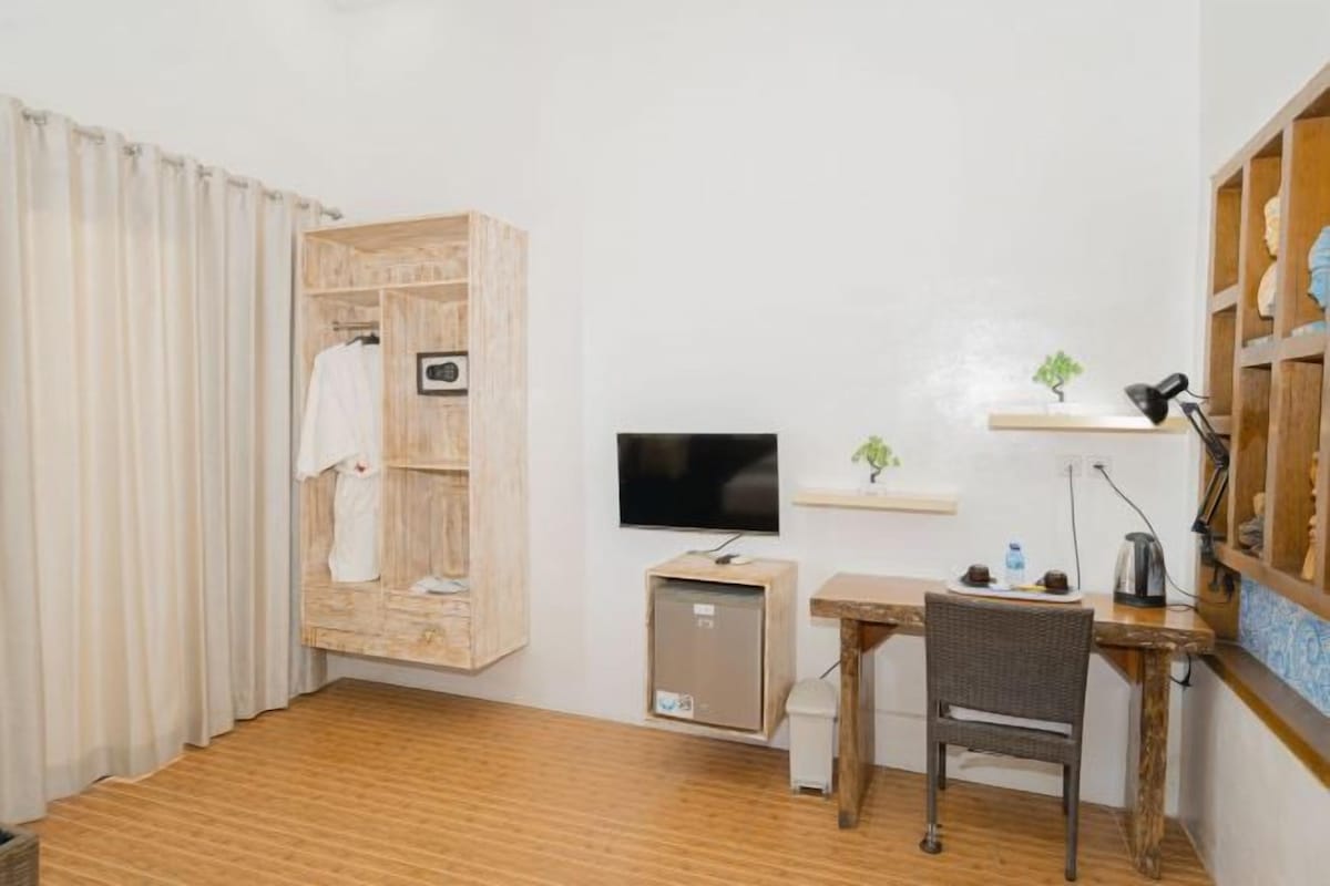 A hotel room with a wooden wardrobe, a television, a small desk with a chair, and a shelf with decorative items.