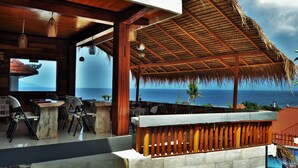 An outdoor dining area with a view of the ocean, wooden furniture, and a thatched roof structure. - Pandawa Beach Resort and Spa Luxury (Penida Island)