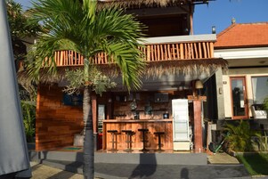 Una zona de bar con barra de madera, techo de paja, taburetes y refrigerador.