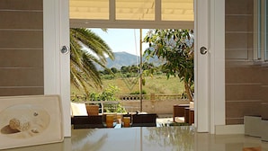 Una habitación con un gran ventanal que da a un jardín y montañas. Hay un cuadro en el mostrador y una pequeña mesa con una silla.