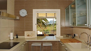 Una cocina moderna con vista a un paisaje tropical a través de un ventanal, un reloj en la pared y un taburete de bar.