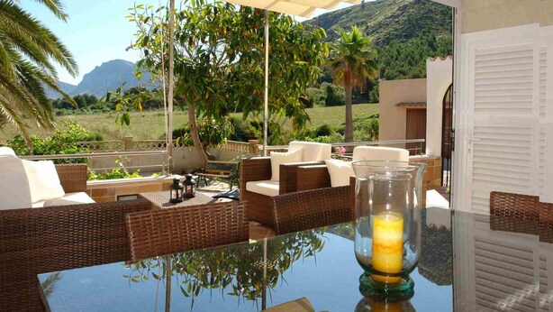 Un espacio al aire libre iluminado por el sol con muebles de mimbre, una mesa de vidrio y una vela.